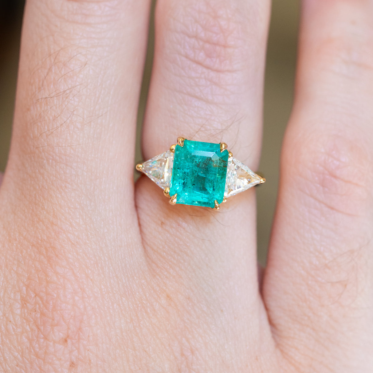 Emerald and diamond three stone ring with emerald cut centre and trilliant diamond side stones close up on hand.