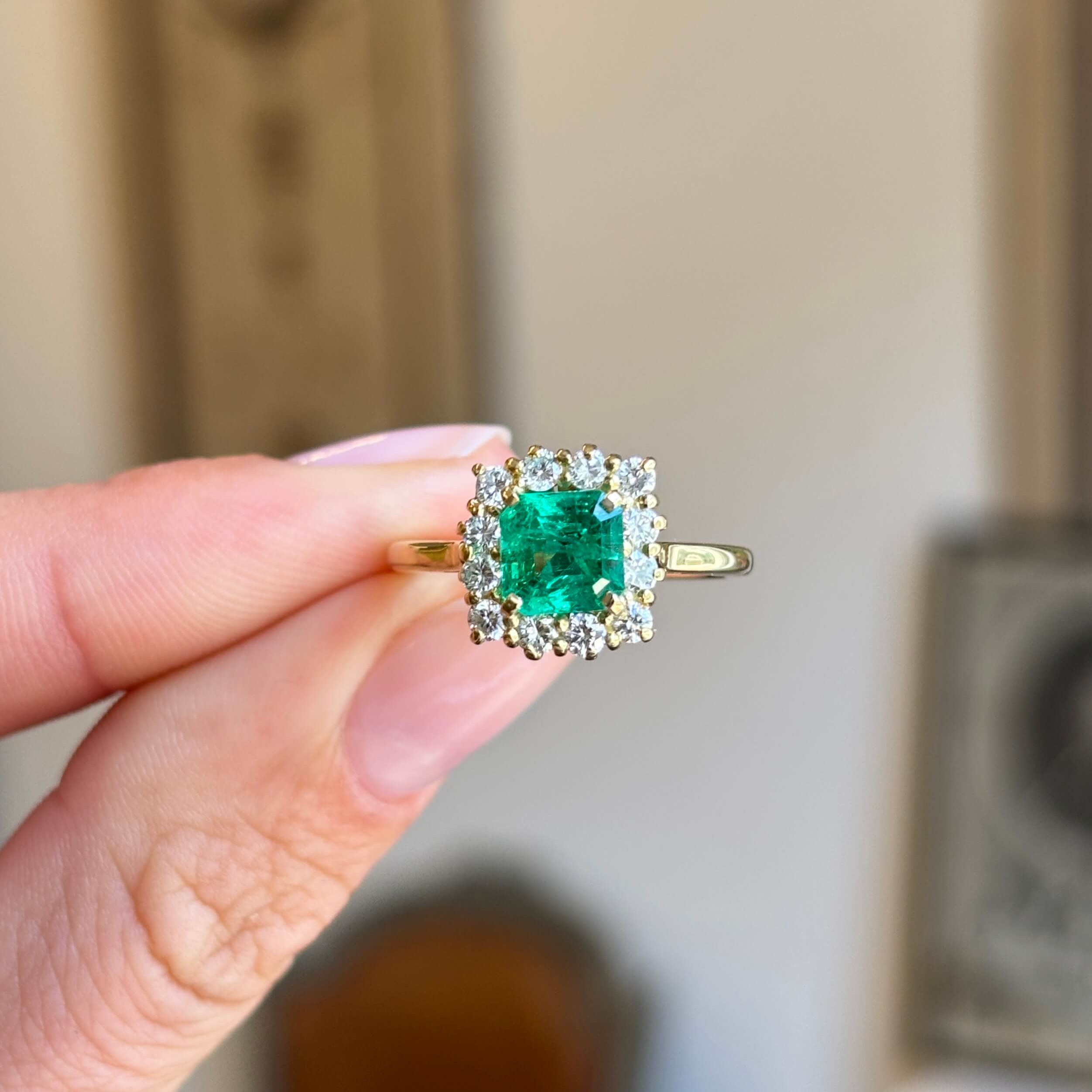 Vintage 1980s Colombian emerald and diamond cluster engagement ring featuring an emerald-cut Colombian emerald surrounded by a halo of brilliant-cut diamonds, held by hand.
