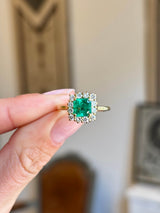 Vintage 1980s Colombian emerald and diamond cluster engagement ring featuring an emerald-cut Colombian emerald surrounded by a halo of brilliant-cut diamonds, held by hand.
