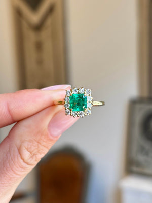 Vintage 1980s Colombian emerald and diamond cluster engagement ring featuring an emerald-cut Colombian emerald surrounded by a halo of brilliant-cut diamonds, held by hand.