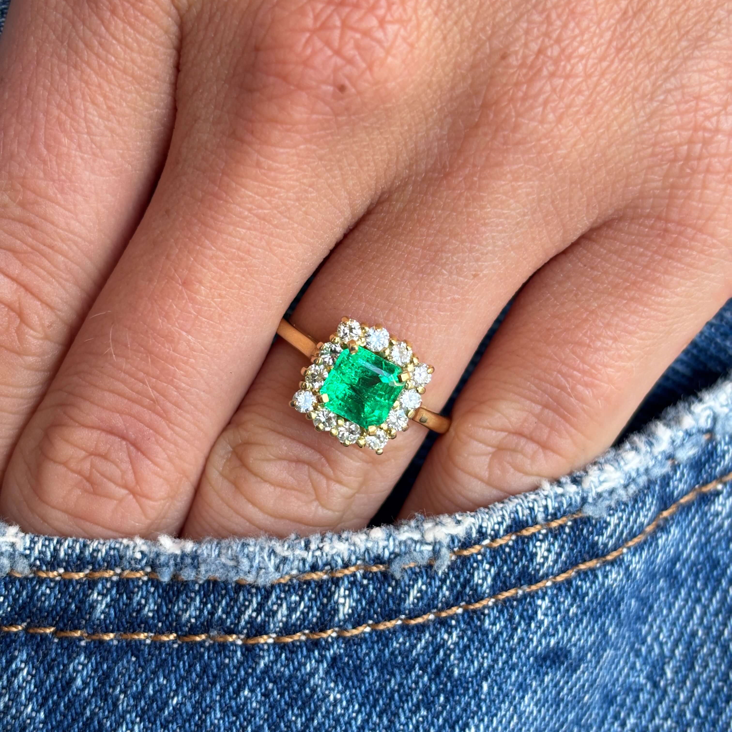 Vintage 1980s Colombian emerald and diamond cluster engagement ring with an emerald-cut Colombian emerald and sparkling diamond halo, worn on a hand on a blue denim background.