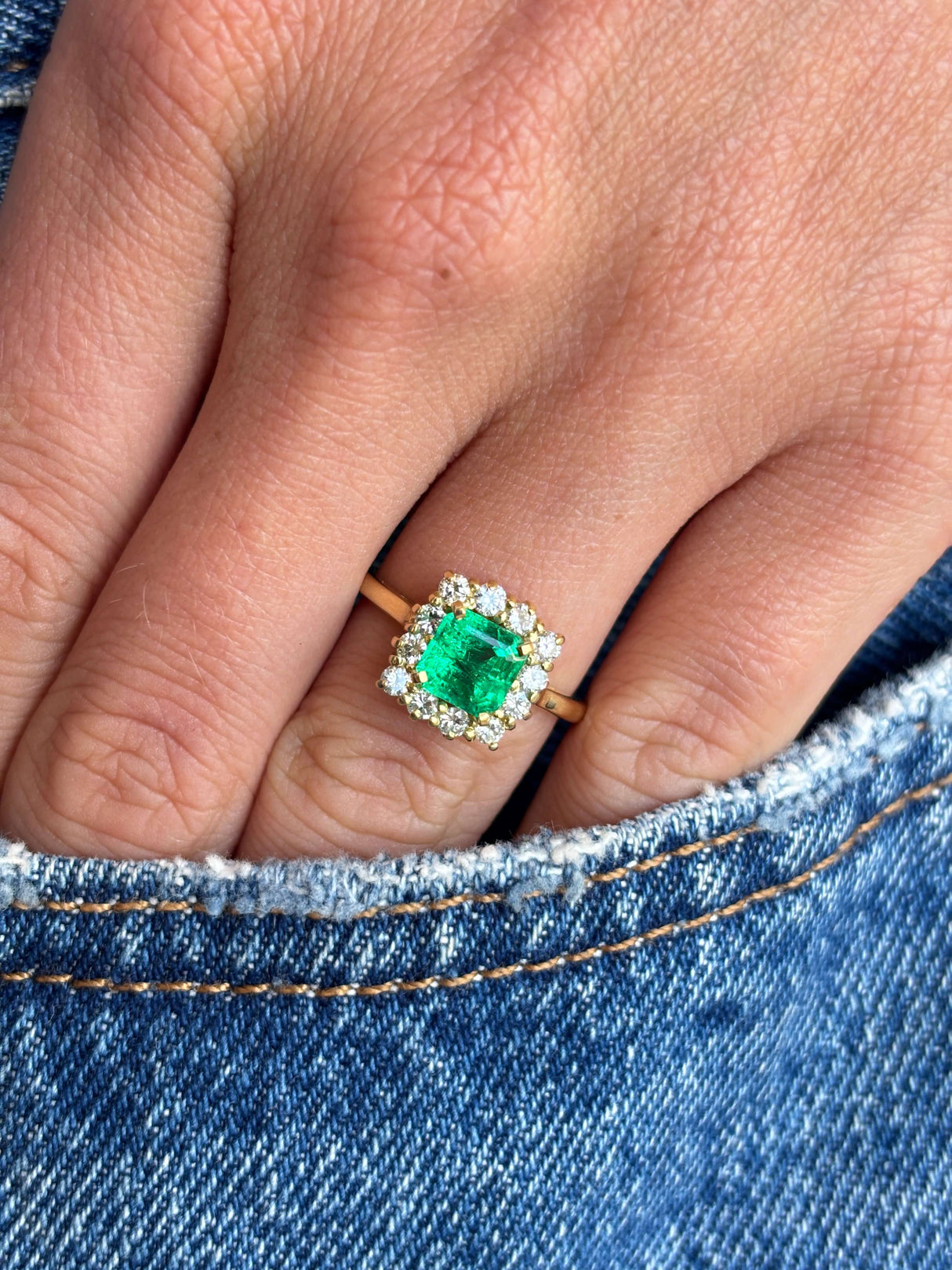 Vintage 1980s Colombian emerald and diamond cluster engagement ring with an emerald-cut Colombian emerald and sparkling diamond halo, worn on a hand on a blue denim background.