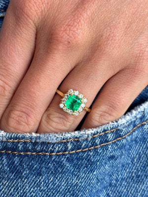 Vintage 1980s Colombian emerald and diamond cluster engagement ring with an emerald-cut Colombian emerald and sparkling diamond halo, worn on a hand on a blue denim background.