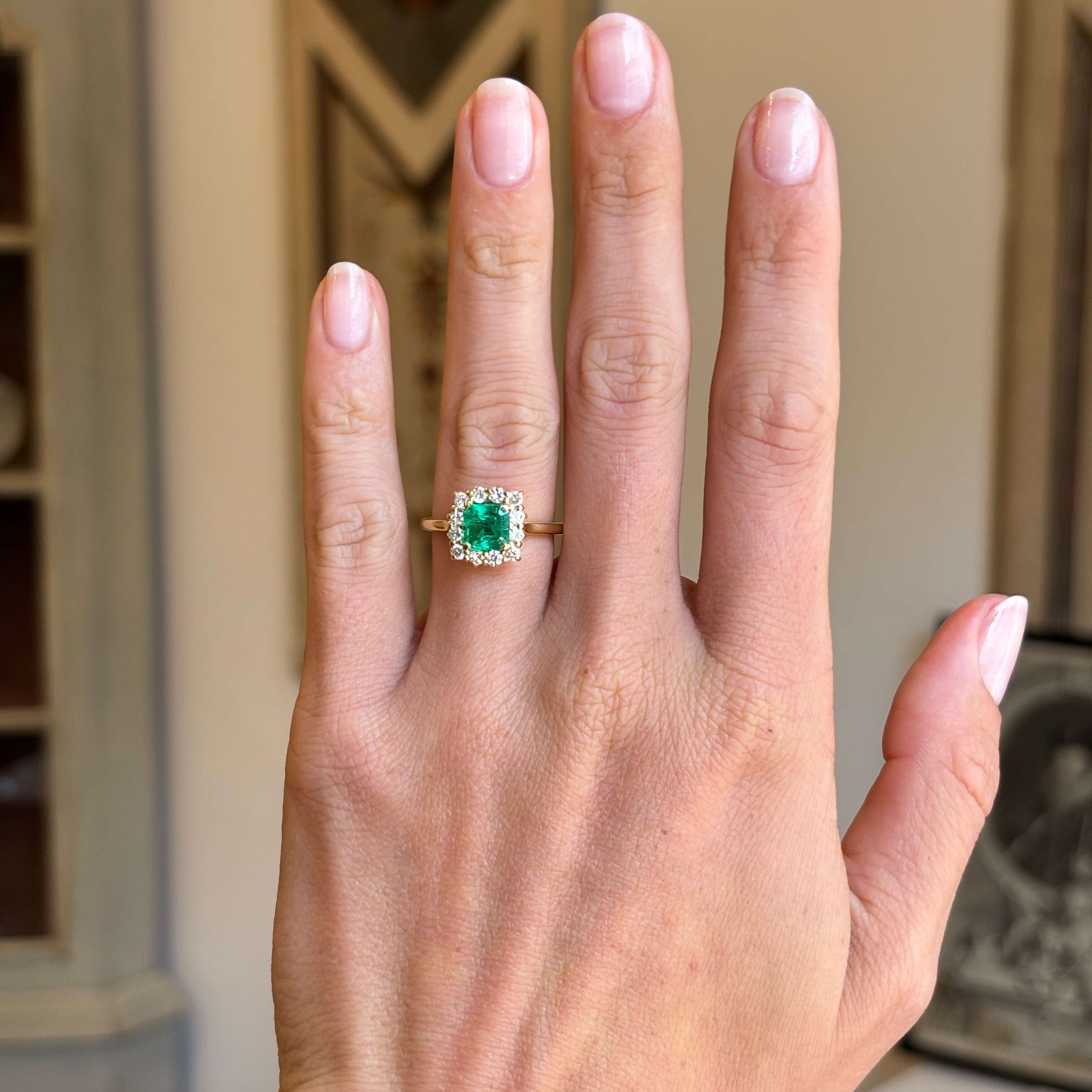 Vintage 1980s Colombian emerald and diamond cluster engagement ring with an emerald-cut Colombian emerald and diamond halo, displayed on a hand.