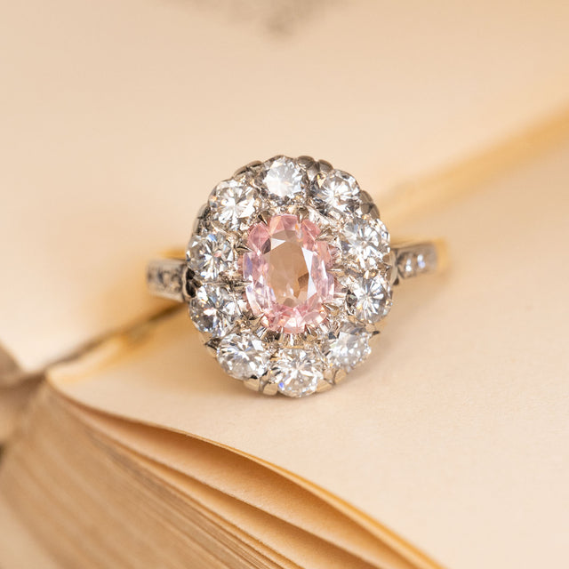 Vintage 2000s cluster ring featuring an orange-pink sapphire surrounded by a halo of brilliant-cut diamonds with diamond-set shoulders.