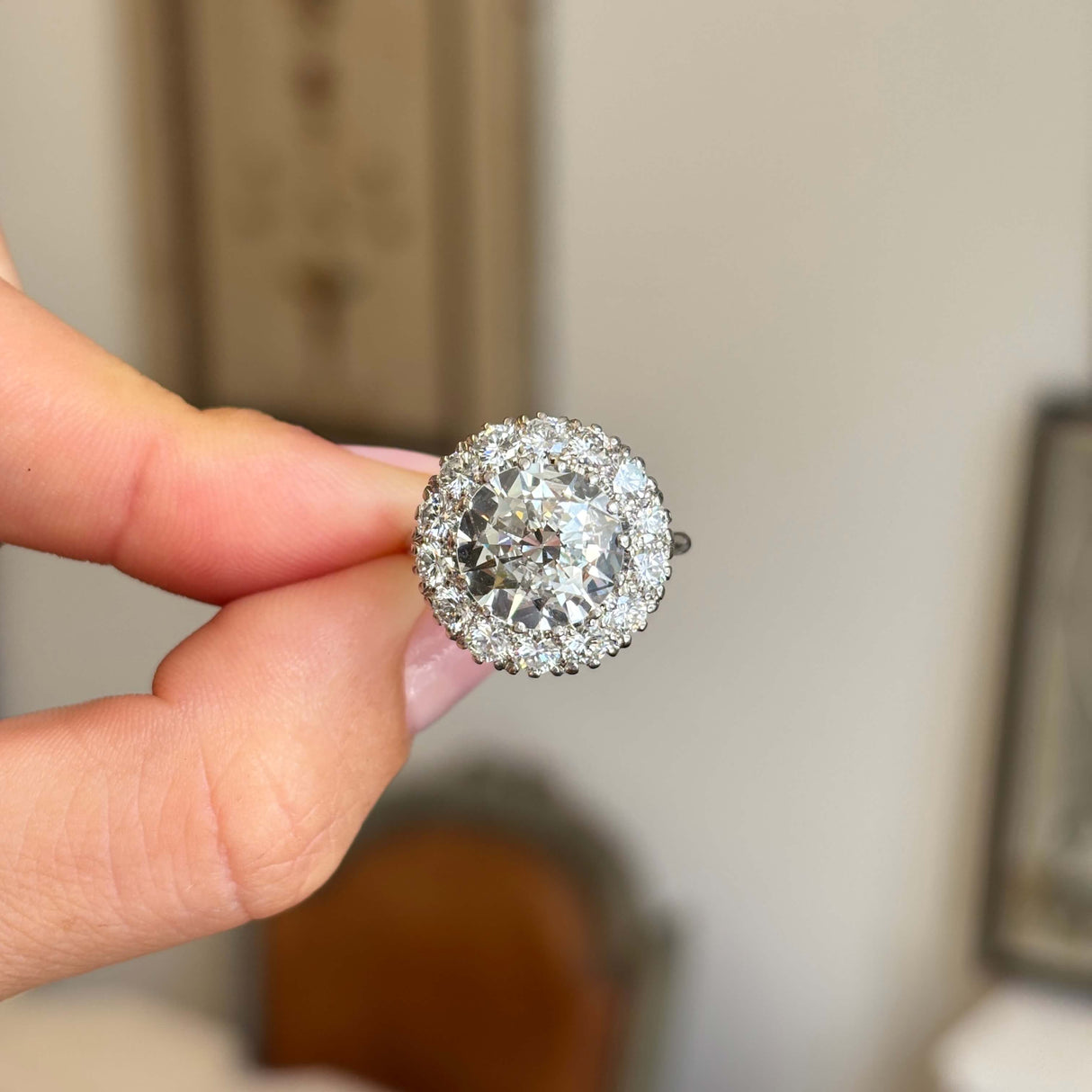 Vintage 1950s platinum diamond cluster engagement ring featuring approximately 4 carats of brilliant diamonds, held between fingers against a softly blurred vintage interior background.