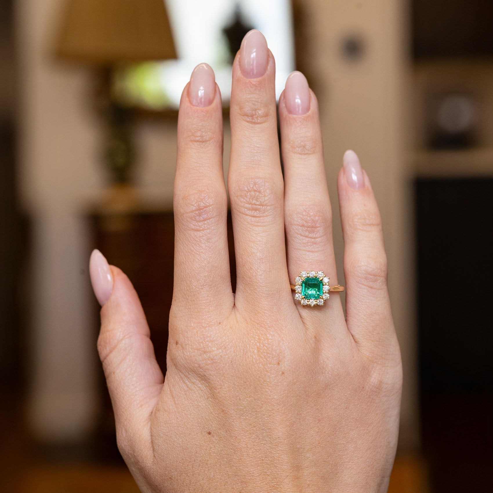 Vintage emerald and diamond cluster engagement ring worn on a hand, showcasing an emerald-cut emerald framed by brilliant-cut diamonds.