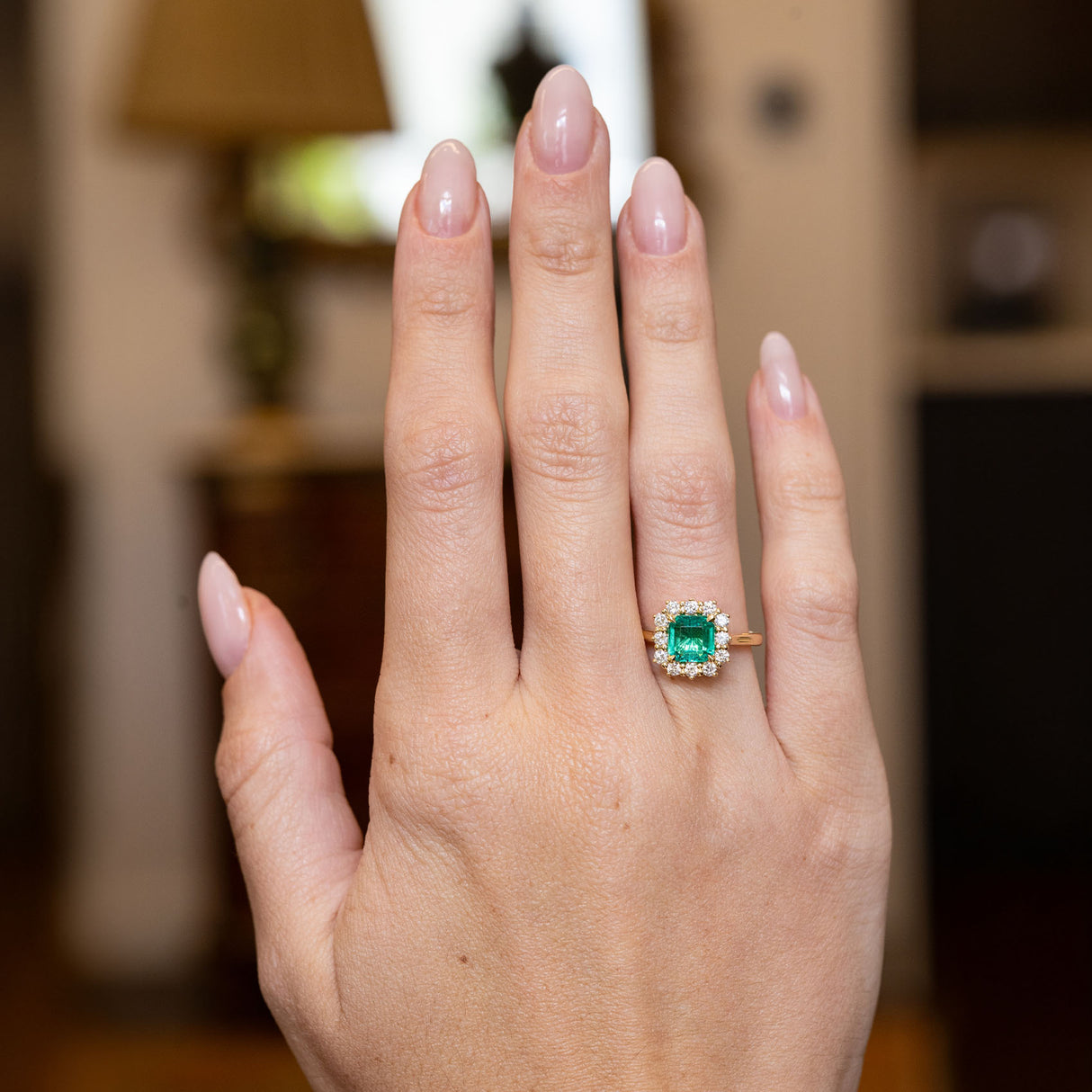 Vintage emerald and diamond cluster engagement ring worn on a hand, showcasing an emerald-cut emerald framed by brilliant-cut diamonds.