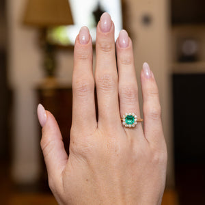 Vintage emerald and diamond cluster engagement ring worn on a hand, showcasing an emerald-cut emerald framed by brilliant-cut diamonds.