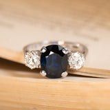 Vintage 1970s midnight-blue sapphire and diamond three-stone ring featuring an oval-cut deep blue sapphire flanked by round brilliant-cut diamonds in white gold.