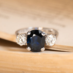 Vintage 1970s midnight-blue sapphire and diamond three-stone ring featuring an oval-cut deep blue sapphire flanked by round brilliant-cut diamonds in white gold.