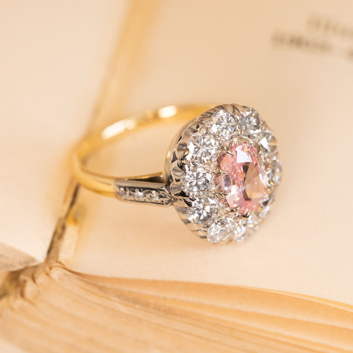 Vintage engagement ring featuring an orange-pink sapphire encircled by brilliant diamonds with elegant diamond-set shoulders and intricate gallery detailing.
