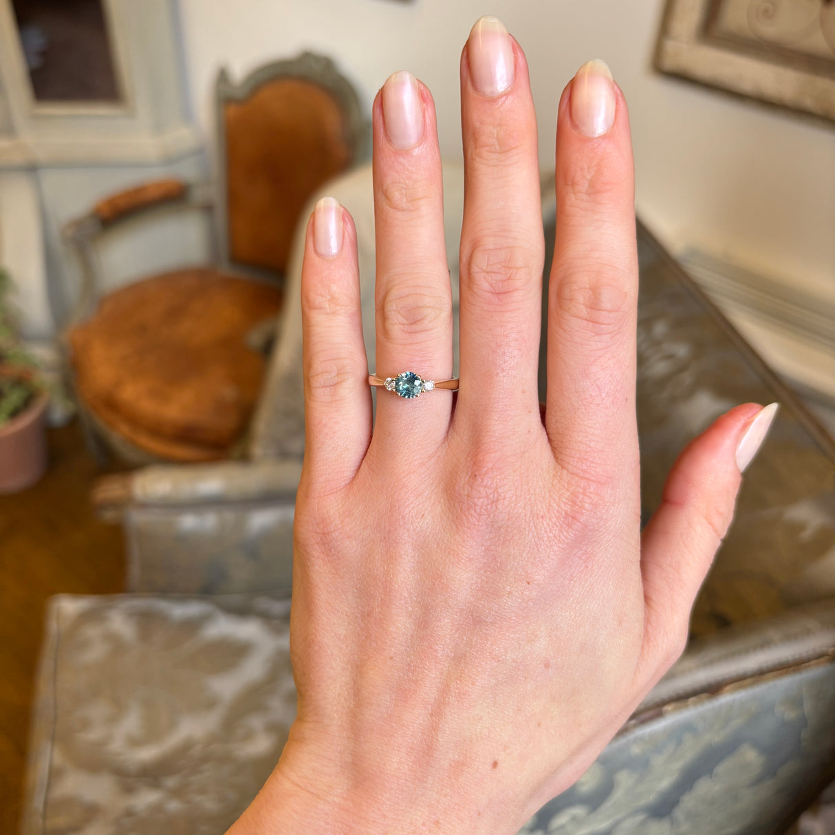 Yellow gold engagement ring with round teal-green sapphire and diamond side stones worn on hand.